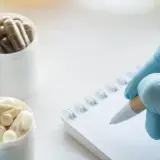 Blaue Handschuhe halten eine Kapsel, daneben stehen Behälter mit Pillen. Eine Hand mit Handschuh schreibt auf einem Notizbuch. Ein Einblick in das Oecotrophologie Studium.