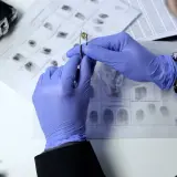 Ein Experte im digitalen forensik studium untersucht eine Patrone mit einer Pinzette, während Fingerabdrücke auf einem Blatt Papier liegen.
