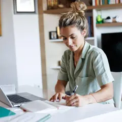 Junge Frau schreibt Notizen, während sie am Laptop arbeitet. Sie plant ihre nächsten Schritte, um ihren Dolmetscher Verdienst zu steigern.