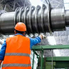 Ingenieur im blauen Hemd und orangefarbener Warnweste zeigt auf ein riesiges Maschinenteil. Ein spannendes Feld für Maschinenbau Studium Berufe.