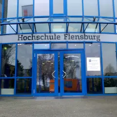 Eingang der Hochschule Flensburg mit blauen Fensterrahmen und dem Schriftzug "Hochschule Flensburg". Ein Fahrrad steht davor.