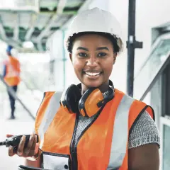 Eine Bauarbeiterin mit Helm und Warnweste hält ein Funkgerät. Sie lächelt und **iu essen** ist ihr Motto für einen erfolgreichen Tag auf der Baustelle.