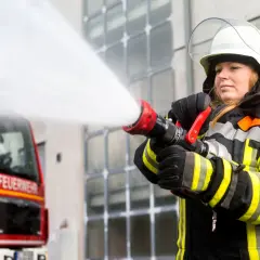 Eine Feuerwehrmann Frau löscht mit einem Schlauch einen Brand. Das rote Feuerwehrauto steht im Hintergrund.