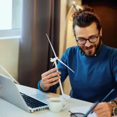 Ein Mann mit Brille und Bart hält ein Modell einer Windkraftanlage. Er studiert es, während er Notizen macht. Ein Laptop und eine Pflanze stehen auf dem Tisch. Ein angehender Wirtschaftsingenieur im Bereich erneuerbare Energien.