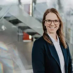 Lächelnde Frau mit Brille und Blazer vor moderner Glasfassade, inspiriert vom Studium.