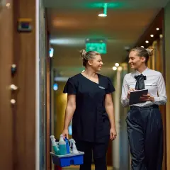 Zwei Frauen im Hotelgang, eine mit Reinigungsmitteln, die andere mit Tablet. Sie sprechen über das Hauswirtschafterin Gehalt.