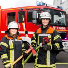 Zwei Feuerwehrleute mit Helm und Schutzkleidung stehen vor einem roten Feuerwehrauto. Sie halten Werkzeuge bereit. So kommt man zur Berufsfeuerwehr.