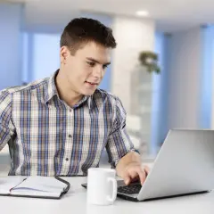 Junger Mann am Laptop, Notizen machend. Er informiert sich über das kaufmann e commerce gehalt.