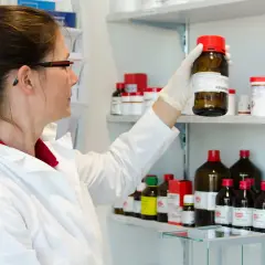 A woman in a lab coat examines a bottle on a shelf filled with chemicals. She's preparing for her state exam, wondering wie lange dauert ein staatsexamen.