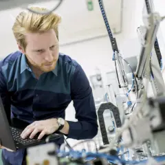 Ein junger ingenieur mit Bart arbeitet konzentriert an einem komplexen mechanischen Aufbau und bedient dabei einen Laptop.