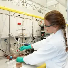 Junge Frau im Labor mit Schutzbrille und Handschuhen, die an einem Experiment arbeitet. Ein Schritt auf dem Weg zum Promotion Studium.