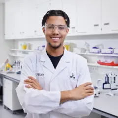 Ein junger Mann im Laborkittel und Schutzbrille, mit verschränkten Armen lächelnd. Er hat seinen Traumjob gefunden: mein traumberuf im Labor.