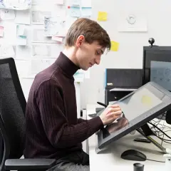 Ein junger Mann im Rollkragenpullover arbeitet konzentriert an seinem Zeichentablett. An der Wand hängen Skizzen und Notizen, die auf sein **Studium Animation** hindeuten.