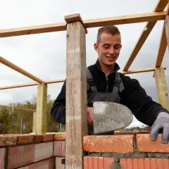 Ein junger Maurer mit Mörtel auf seiner Kelle baut eine Ziegelwand. Er lächelt, während er arbeitet. Die Frage "wie viel verdient man als maurer" beschäftigt viele.