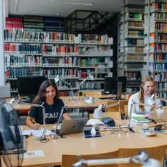 Zwei Studentinnen lernen in einer Bibliothek. Die Bedeutung von Bildung wird hier durch konzentriertes Arbeiten an Laptops und Büchern promoviert.