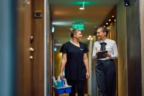 Zwei Frauen im Hotelgang, eine mit Reinigungsmitteln, die andere mit Tablet. Sie sprechen über das Hauswirtschafterin Gehalt.