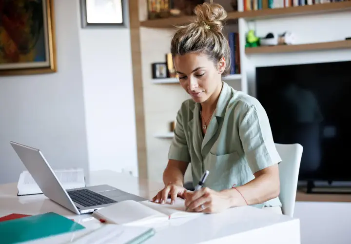Junge Frau schreibt Notizen, während sie am Laptop arbeitet. Sie plant ihre nächsten Schritte, um ihren Dolmetscher Verdienst zu steigern.