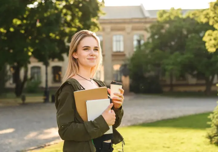 Junge Frau mit Büchern und Kaffee vor einem Universitätsgebäude. Sie denkt darüber nach, wie lange dauert ein Masterstudium.