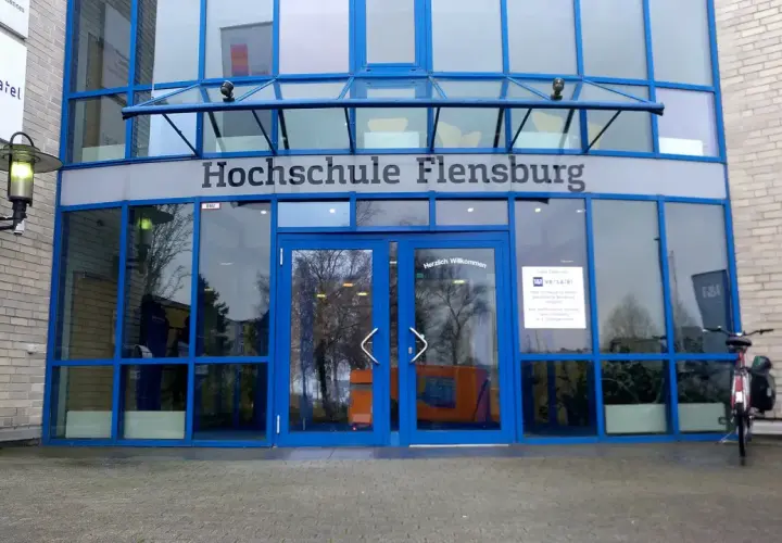 Eingang der Hochschule Flensburg mit blauen Fensterrahmen und dem Schriftzug "Hochschule Flensburg". Ein Fahrrad steht davor.