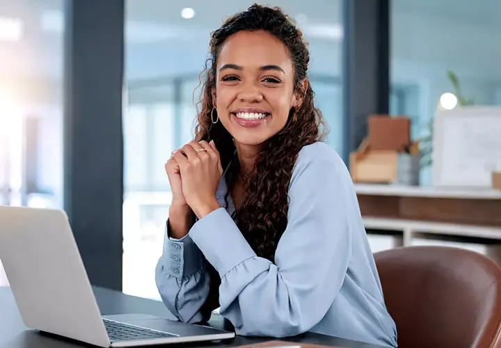 Lächelnde junge Frau am Schreibtisch mit Laptop. Sie informiert sich über den industriekaufmann verdienst ausbildung.