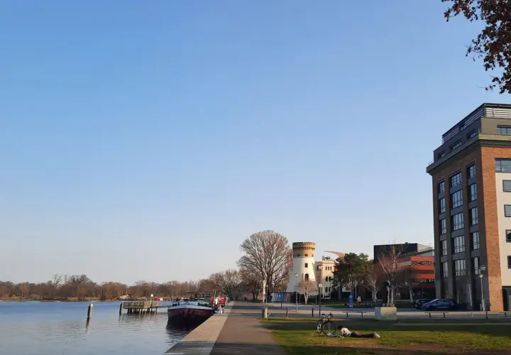 Ein sonniger Tag am Wasser in Potsdam. Ein Mann liest im Gras, ein Fahrrad steht daneben. Im Hintergrund ein Turm und moderne Gebäude.