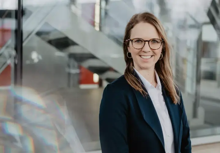 Lächelnde Frau mit Brille und Blazer vor moderner Glasfassade, inspiriert vom Studium.