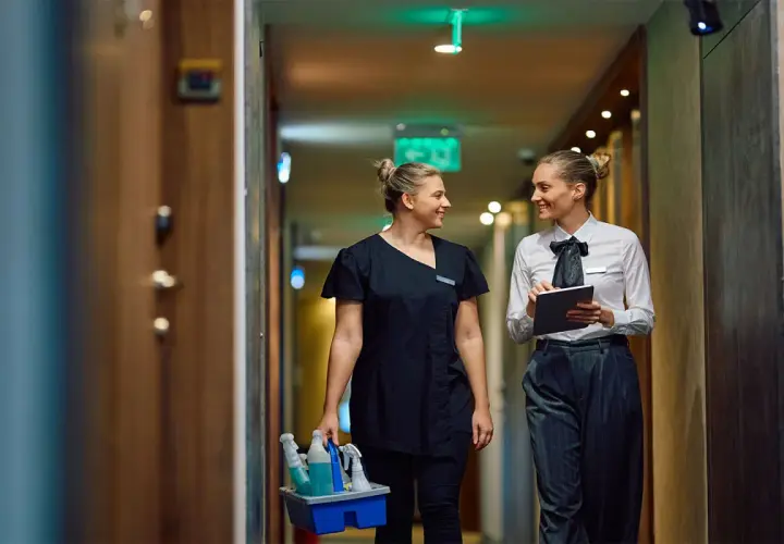 Zwei Frauen im Hotelgang, eine mit Reinigungsmitteln, die andere mit Tablet. Sie sprechen über das Hauswirtschafterin Gehalt.