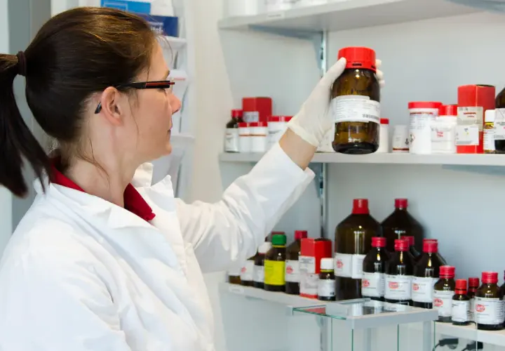 A woman in a lab coat examines a bottle on a shelf filled with chemicals. She's preparing for her state exam, wondering wie lange dauert ein staatsexamen.