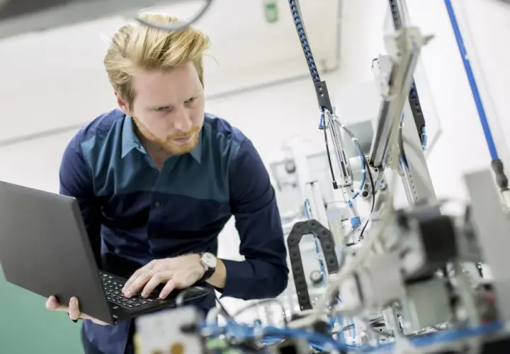 Ein junger ingenieur mit Bart arbeitet konzentriert an einem komplexen mechanischen Aufbau und bedient dabei einen Laptop.