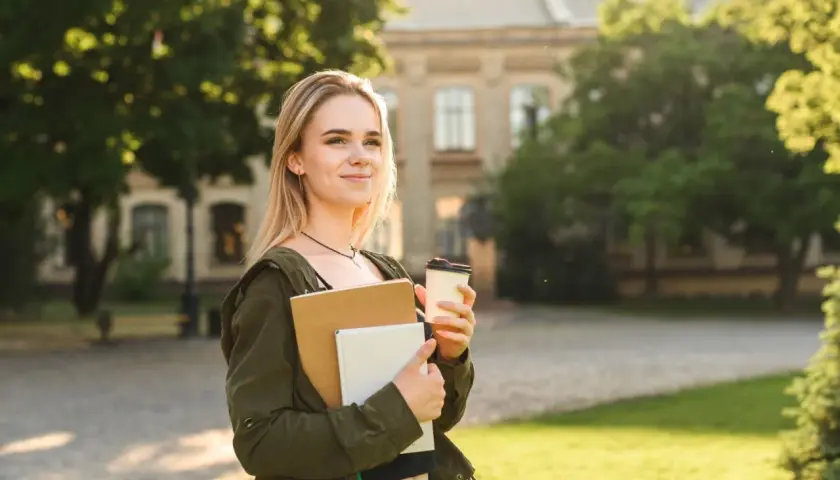 Junge Frau mit Büchern und Kaffee vor einem Universitätsgebäude. Sie denkt darüber nach, wie lange dauert ein Masterstudium.