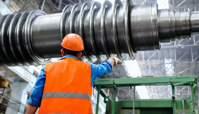 Ingenieur im blauen Hemd und orangefarbener Warnweste zeigt auf ein riesiges Maschinenteil. Ein spannendes Feld für Maschinenbau Studium Berufe.