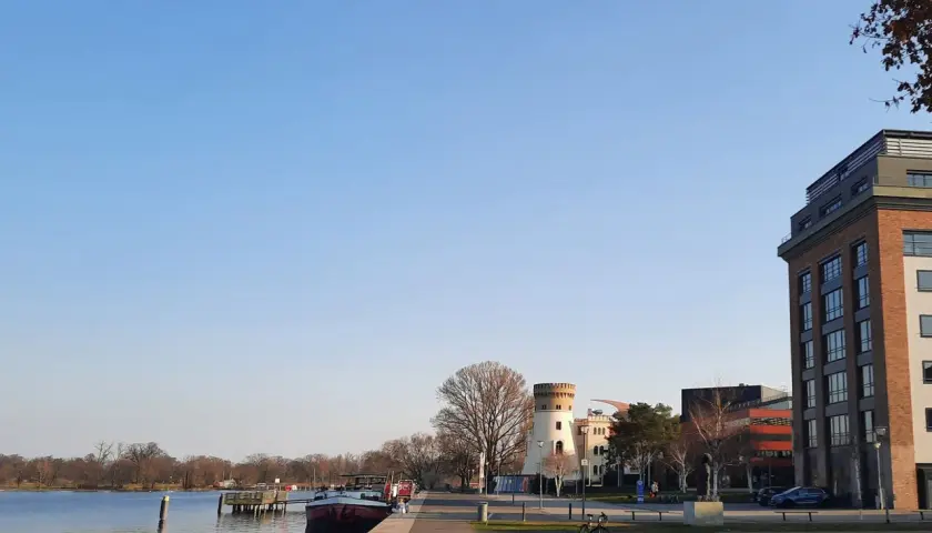 Ein sonniger Tag am Wasser in Potsdam. Ein Mann liest im Gras, ein Fahrrad steht daneben. Im Hintergrund ein Turm und moderne Gebäude.