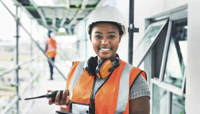Eine Bauarbeiterin mit Helm und Warnweste hält ein Funkgerät. Sie lächelt und **iu essen** ist ihr Motto für einen erfolgreichen Tag auf der Baustelle.