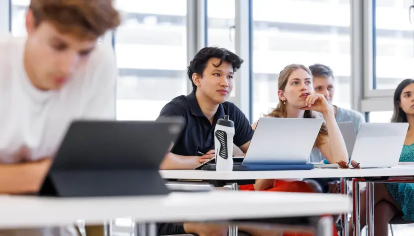 Studenten im Hörsaal, die sich auf ihre Lektionen konzentrieren. Ein junger Mann mit einem schwarzen Poloshirt und einer Wasserflasche, der aufmerksam zuhört. Dies ist ein typisches Bild für Management and Technology TUM.