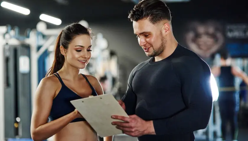 Ein Trainer bespricht mit einer Kundin Trainingspläne. Das Gehalt eines Sport- und Fitnesskaufmanns nach der Ausbildung ist attraktiv.