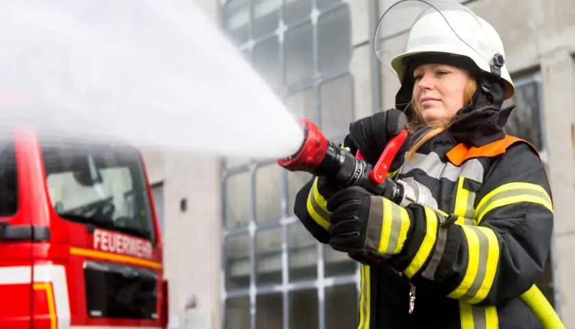 Eine Feuerwehrmann Frau löscht mit einem Schlauch einen Brand. Das rote Feuerwehrauto steht im Hintergrund.