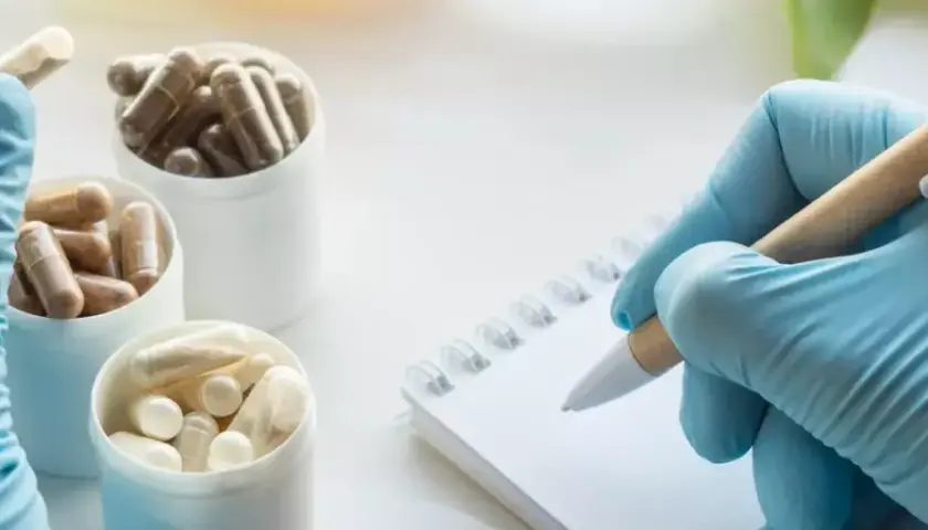 Blaue Handschuhe halten eine Kapsel, daneben stehen Behälter mit Pillen. Eine Hand mit Handschuh schreibt auf einem Notizbuch. Ein Einblick in das Oecotrophologie Studium.