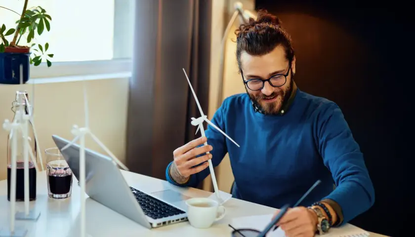 Ein Mann mit Brille und Bart hält ein Modell einer Windkraftanlage. Er studiert es, während er Notizen macht. Ein Laptop und eine Pflanze stehen auf dem Tisch. Ein angehender Wirtschaftsingenieur im Bereich erneuerbare Energien.