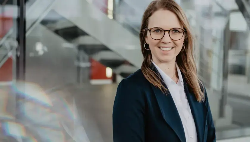 Lächelnde Frau mit Brille und Blazer vor moderner Glasfassade, inspiriert vom Studium.