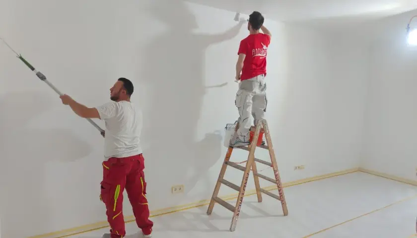 Zwei Maler streichen Wände und Decke weiß. Die Ausbildung zum Maler und Lackierer bietet ein gutes Gehalt netto.