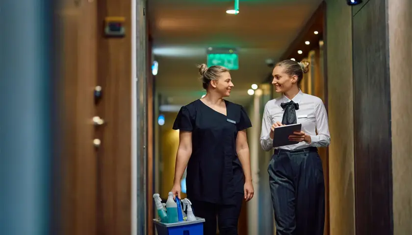 Zwei Frauen im Hotelgang, eine mit Reinigungsmitteln, die andere mit Tablet. Sie sprechen über das Hauswirtschafterin Gehalt.