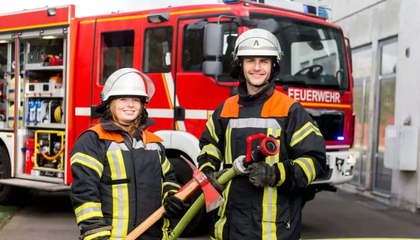 Zwei Feuerwehrleute mit Helm und Schutzkleidung stehen vor einem roten Feuerwehrauto. Sie halten Werkzeuge bereit. So kommt man zur Berufsfeuerwehr.