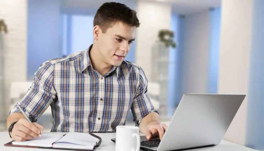 Junger Mann am Laptop, Notizen machend. Er informiert sich über das kaufmann e commerce gehalt.
