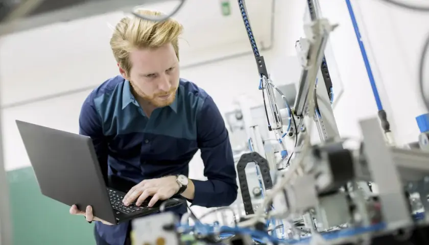 Ein junger ingenieur mit Bart arbeitet konzentriert an einem komplexen mechanischen Aufbau und bedient dabei einen Laptop.