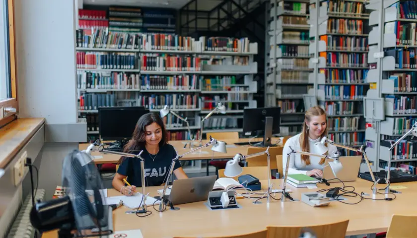 Zwei Studentinnen lernen in einer Bibliothek. Die Bedeutung von Bildung wird hier durch konzentriertes Arbeiten an Laptops und Büchern promoviert.