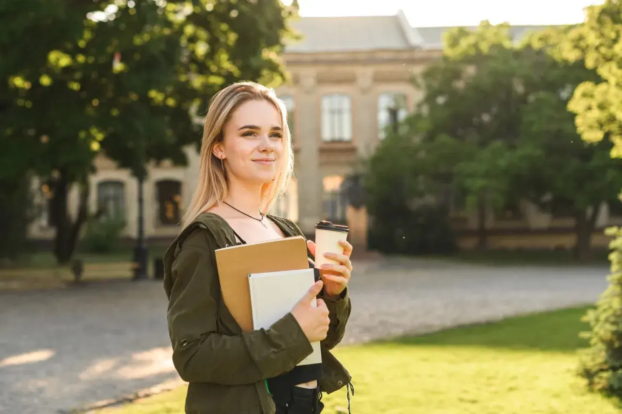 Junge Frau mit Büchern und Kaffee vor einem Universitätsgebäude. Sie denkt darüber nach, wie lange dauert ein Masterstudium.