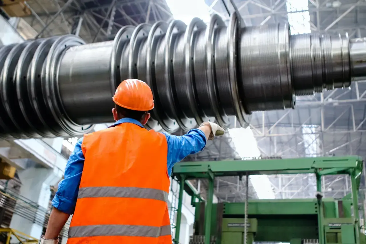 Ingenieur im blauen Hemd und orangefarbener Warnweste zeigt auf ein riesiges Maschinenteil. Ein spannendes Feld für Maschinenbau Studium Berufe.