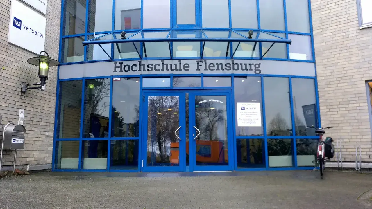 Eingang der Hochschule Flensburg mit blauen Fensterrahmen und dem Schriftzug "Hochschule Flensburg". Ein Fahrrad steht davor.