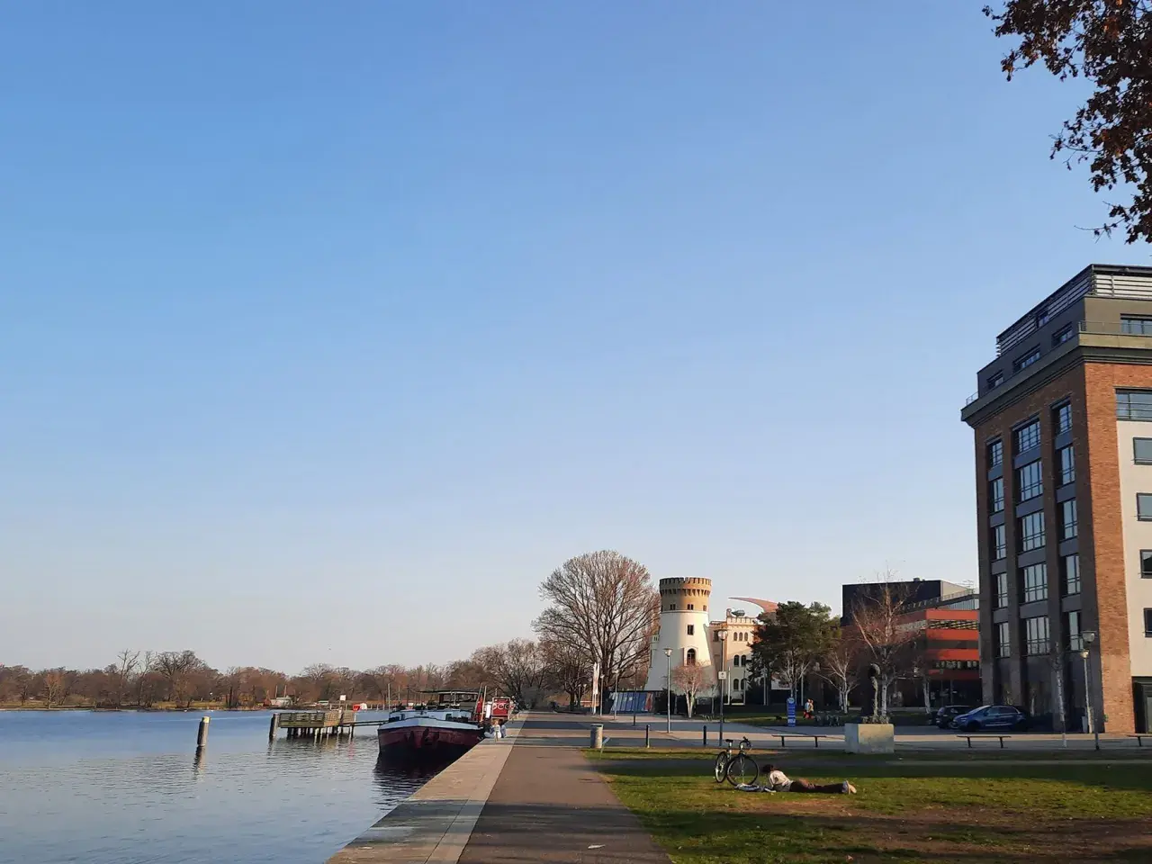 Ein sonniger Tag am Wasser in Potsdam. Ein Mann liest im Gras, ein Fahrrad steht daneben. Im Hintergrund ein Turm und moderne Gebäude.