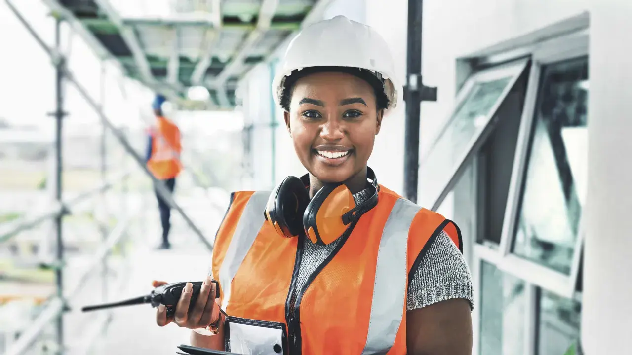 Eine Bauarbeiterin mit Helm und Warnweste hält ein Funkgerät. Sie lächelt und **iu essen** ist ihr Motto für einen erfolgreichen Tag auf der Baustelle.